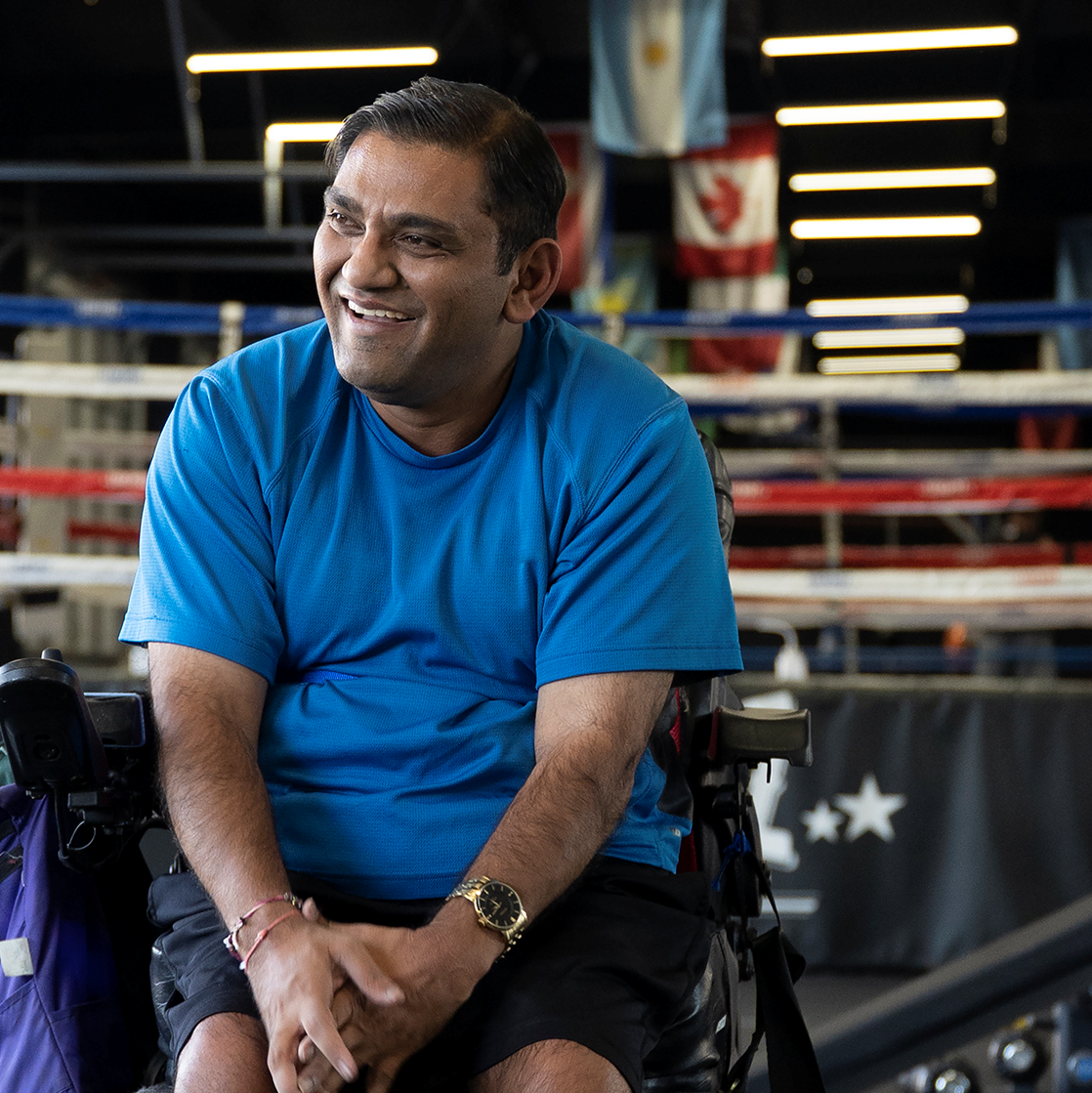 Bhumit_sq A man smiling, with his head turned to the side. He sits in his wheelchair beside a wrestling ring.