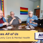 Cover Image for the All of Us Research Program webinar "It Takes All of Us, Community Care and Mental Health". Shows a group of seniors playing cards around a table in a community center. Rainbow flags hang on the wall.
