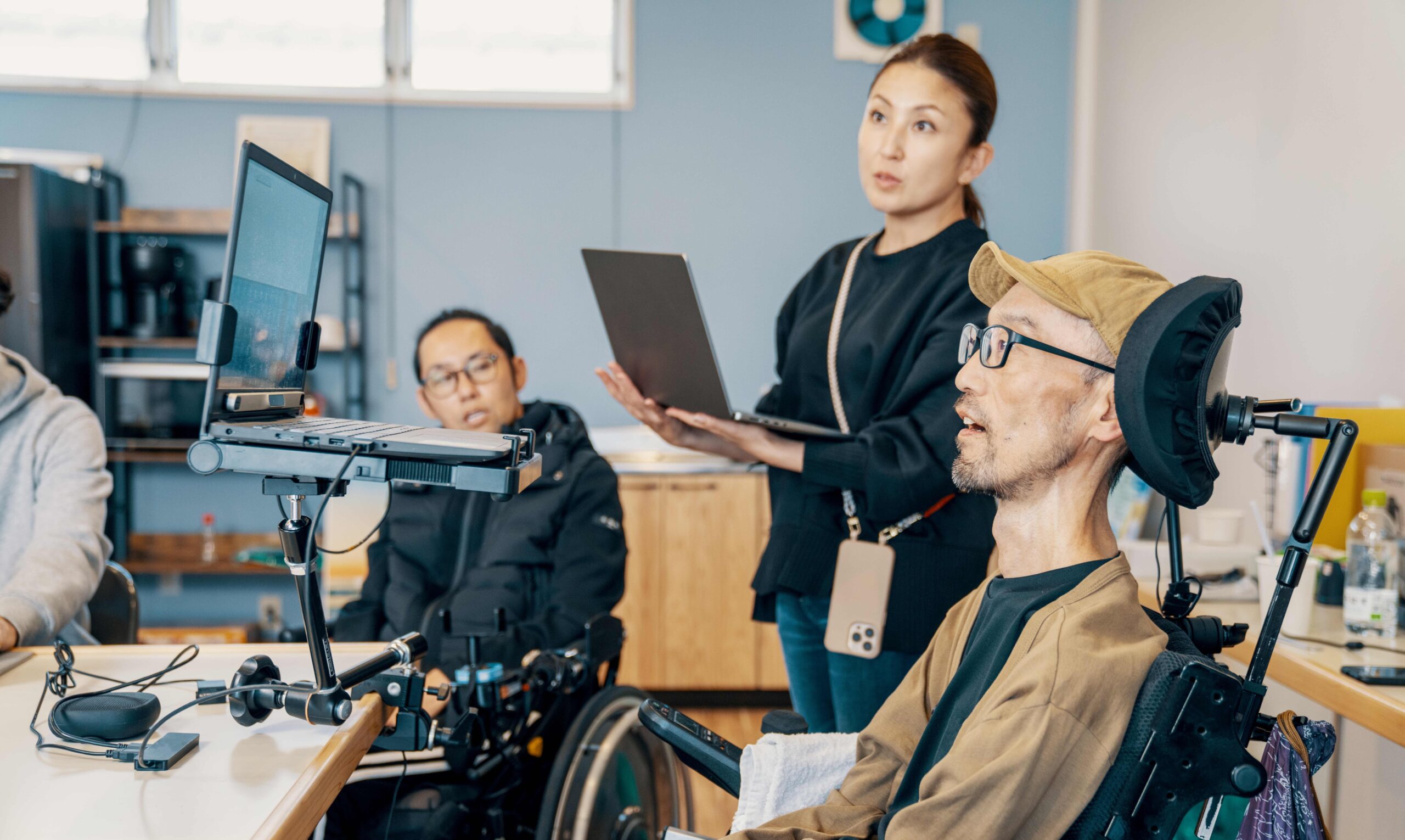 A man in a power wheelchair with a head-rest reviews documents on his laptop, which is on a stand at eye level. He is in a lab with co-workers behind.