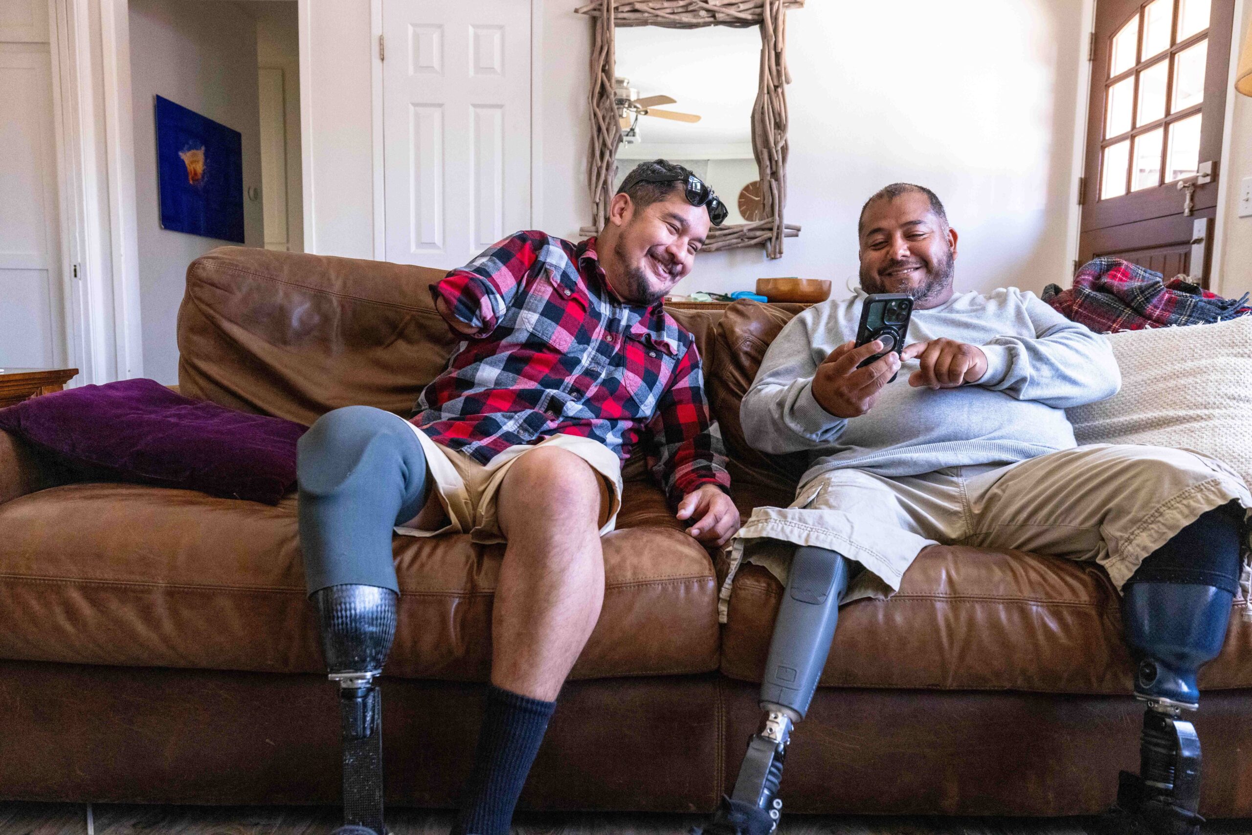 Two men with leg and arm amputations sit together on the couch, laughing as one scrolls through this phone.